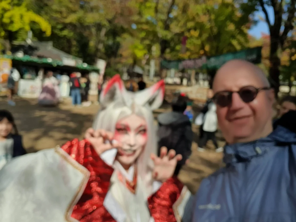 A tourist takes a selfie with a cosplayer dressed as a fox spirit (kitsune) in white and red costume at a Korean outdoor festival.