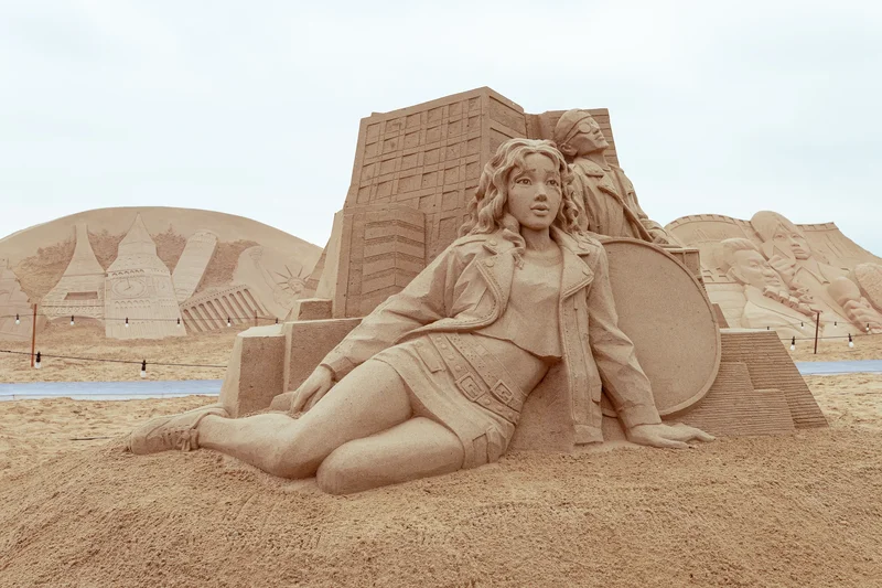 A detailed sand sculpture of a young woman reclining in front of abstract city structures and circular forms, part of the Haeundae Sand Sculpture Festival.